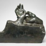 A bronze sculpture of two human hands is shown in front of a white backdrop.