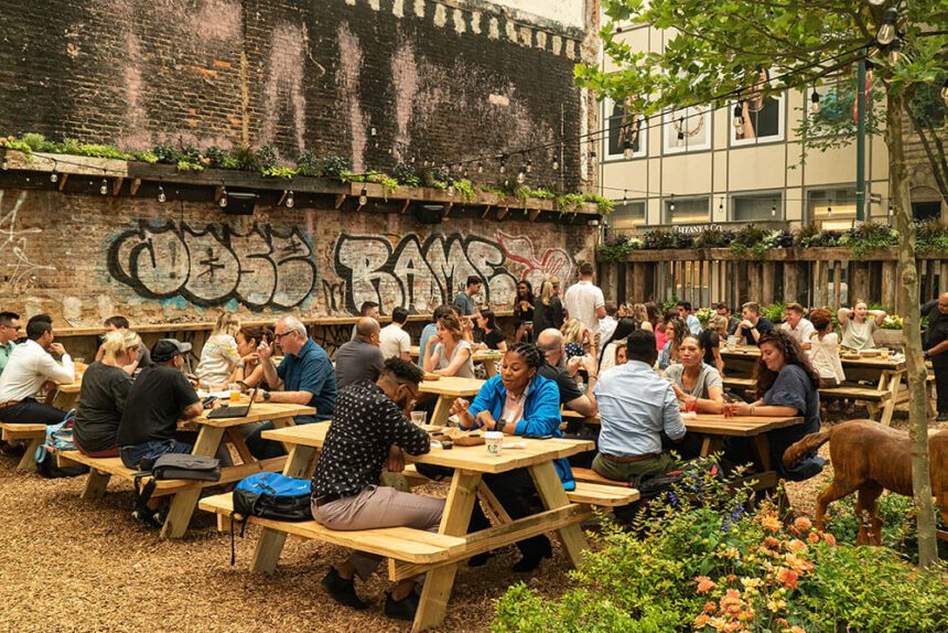 An outdoor beer garden is set up with picnic tables surrounded by lush green plants. There are people shown sitting at the picnic tables. Some are eating, others are drinking. There are lights strung overhead, across the picnic table area.