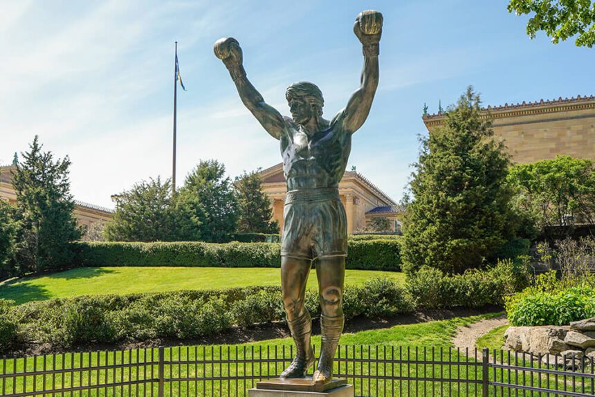 A large statue of a man holding his arms and fists in the air is shown in front of the Philadelphia Museum of Art. There are lush green bushes and lawns behind the statue. The sky above is a bright blue with a few white clouds off to the left. 