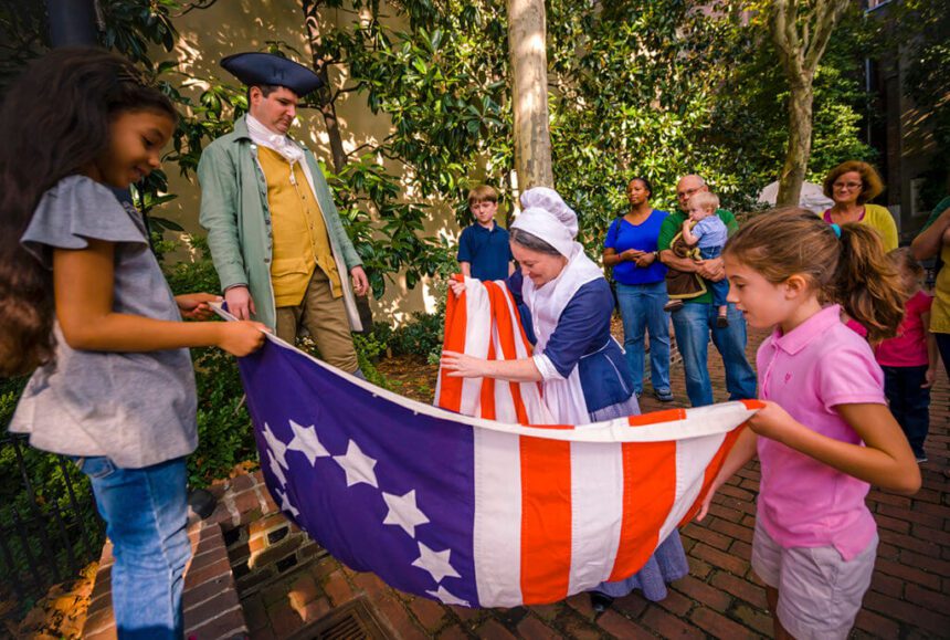 A woman dresses in colonial clothing shows an early American flag to school children.