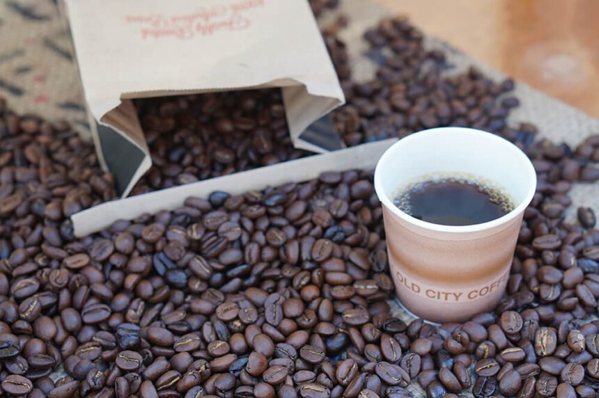 A pile of coffee beans are shown spilled out on a counter. An open bag is off to the back left. A coffee cup filled with coffee is in front to the right.