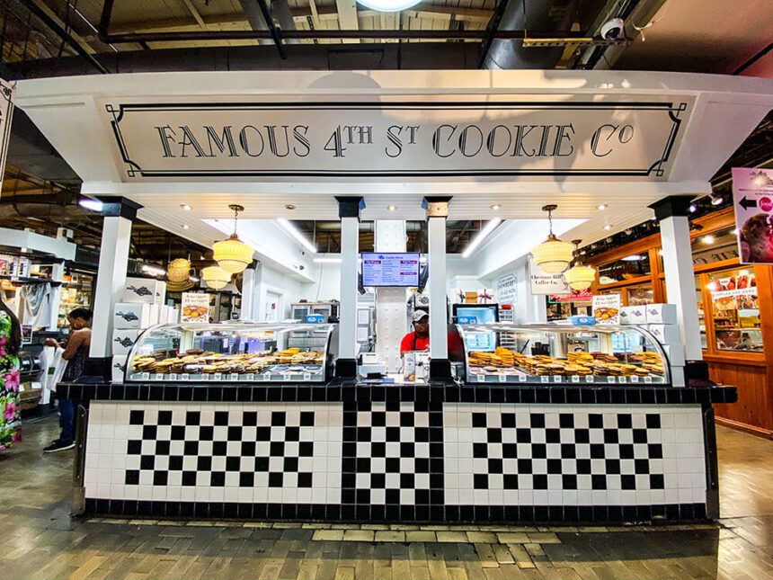 A black and white checkered tile wall lines underneath a counter with cookies in a glass case off to the left and right. A large sign hangs over the stall reading, Famous 4th St Cookie Co