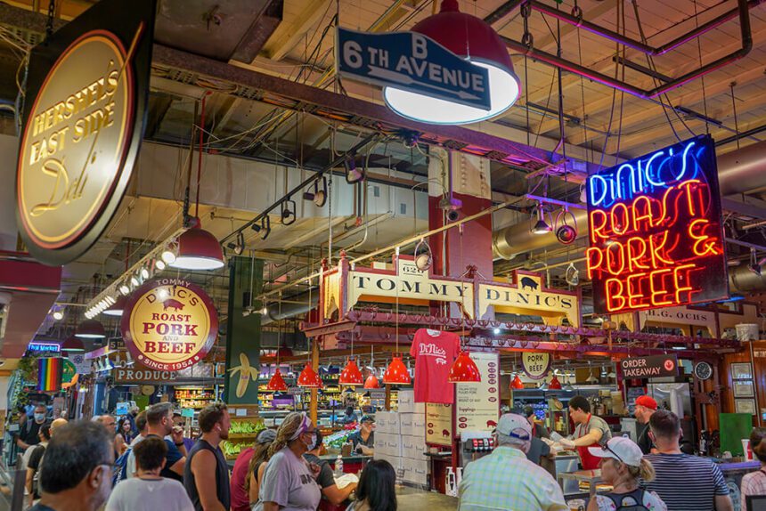 Neon signs hang from the ceiling. One to the right reads DiNic's Roast Pork & Beef. There is a bustle of people surrounding the stall inside of the market. There are bright red lights hanging over the counter where people are eating and ordering meals.