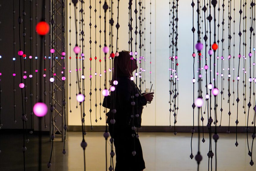 A woman dressed in black is shown walking in a room where there are strands of bright lights hanging as part of an art installation. The lights look like round orbs.