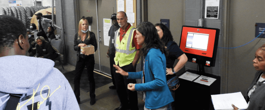 A woman wearing a blue jacket stands in front of a group appearing to offer instructions. People are shown standing around listening. One man behind her wears a bright vest. There is a computer screen shown with a red background.