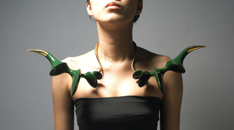A woman is shown wearing an ornate necklace around her neck. The artwork is gold with green horns on the ends pointing outward.