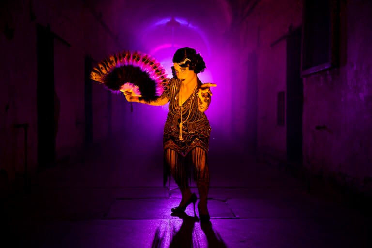 A woman dressed in a flapper costume stands, holding a fan, as she leans forward appearing to coax the camera toward her. There is a purple glow behind her.