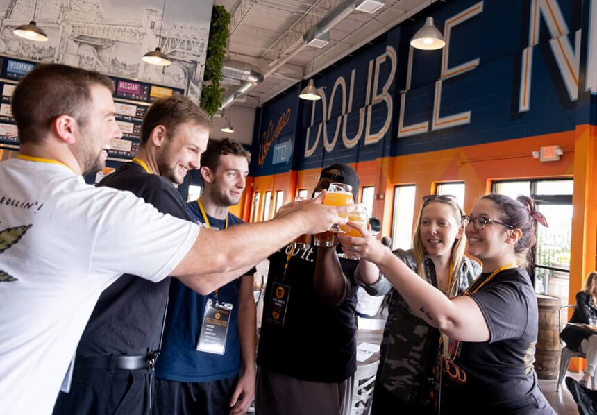 A group of people are shown holding beer glasses in front of them. They are smiling as they clink their glasses together.