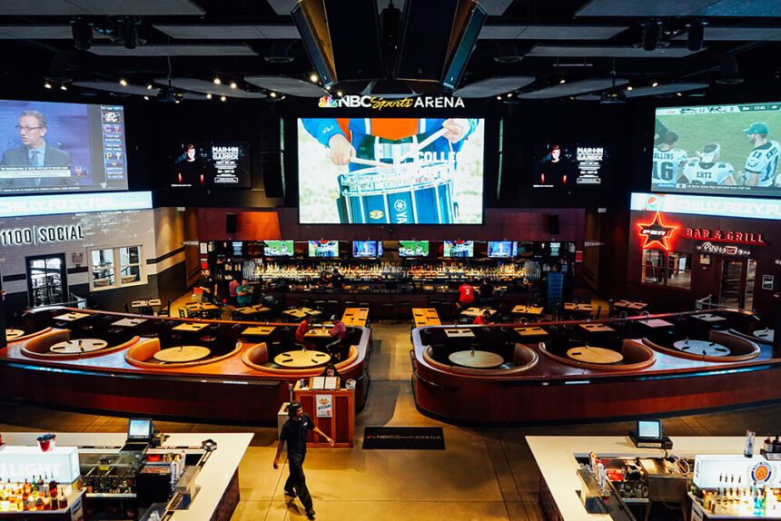 A large, open, indoor space is shown. There are tables and chairs set up, along with booths. There are three very large TV screens hanging over a bar. The bar has an array of bottles set up on shelves behind it. A worker is shown in the forefront, walking past the seating area.