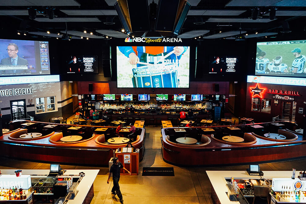 A large, open, indoor space is shown. There are tables and chairs set up, along with booths. There are three very large TV screens hanging over a bar. The bar has an array of bottles set up on shelves behind it. A worker is shown in the forefront, walking past the seating area.