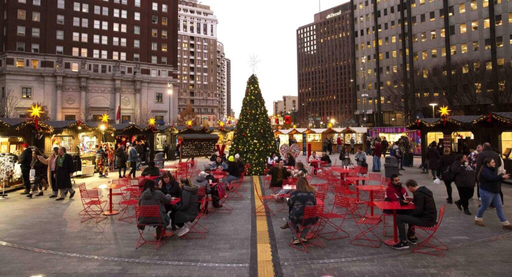 People sitting on red chairs by red tables surrounding a tall holiday tree with white huts around it where vendors are selling goods to customers
