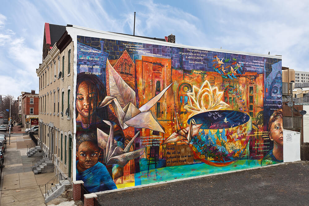 A colorful mural decorates the side of a building. Two young children's faces are on the edge of the mural to the left. Words of a poem are scattered throughout the art. Paper cranes are shown, along with a colorful bowl with smaller paper cranes. Another child's face is off in the right corner of the mural.