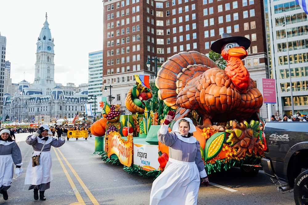 A large float with a turkey wearing a hat rolls down the street. Women dressed as pilgrims walk beside the float and wave. Philadelphia's City Hall is in the background. More people are seen marching behind the float during the annual Thanksgiving Day parade.