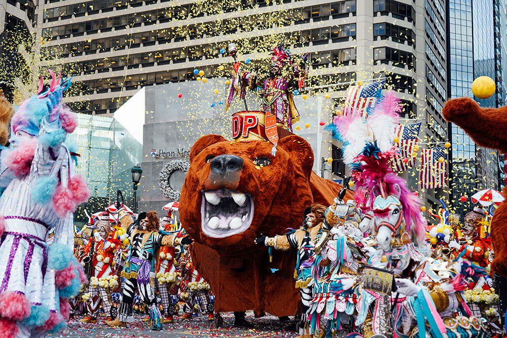 Gold confetti is thrown in the air. A large float to look like a bear is in the middle. A man in a colorful costume stands on top of the float. Other performers in colorful costumes surround the float during the annual Mummers Parade. Some costumes are white, pink, blue, and purple. One performer appears to have a headpiece to look like a horse. American flags hang off of buildings in the background.
