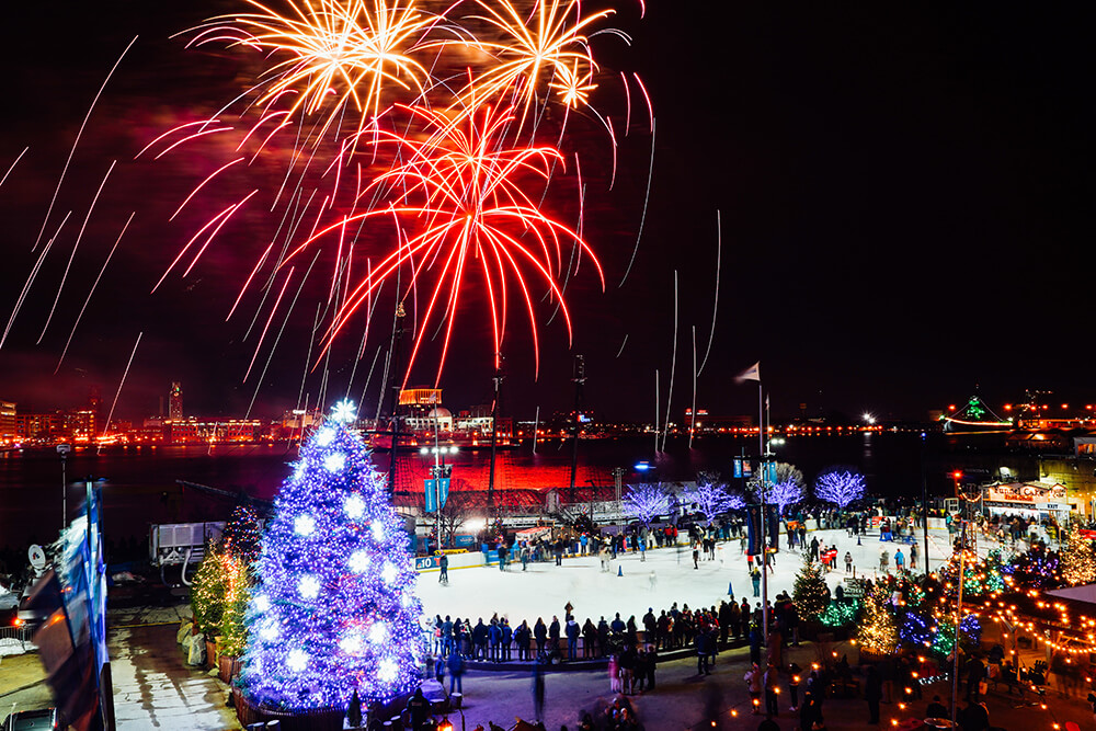 A large tree lights up in purple to the left. Red and gold fireworks soar into the night sky. People gather on an ice rink below, gazing up at the fireworks. Lights are strung nearby the ice rink along with festive winter holiday decorations.