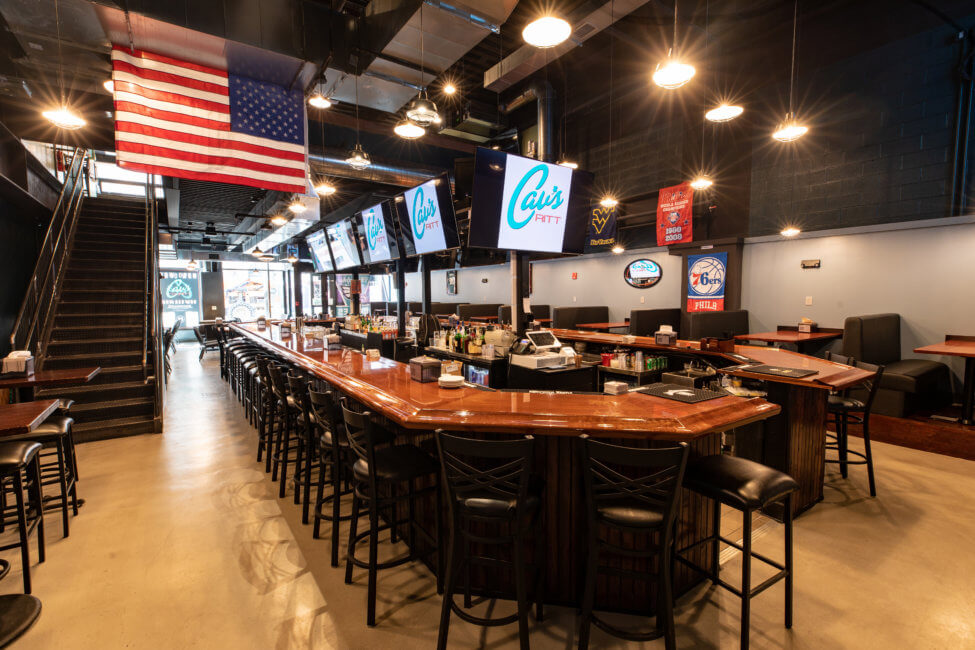 A bar is shown, there are barstools surrounding it. There are several TVs hanging above the bar. A large American flag is hanging from the ceiling. There are open booths off to the right, along the wall. There is a set of stairs leading to the second floor to the left of the bar.