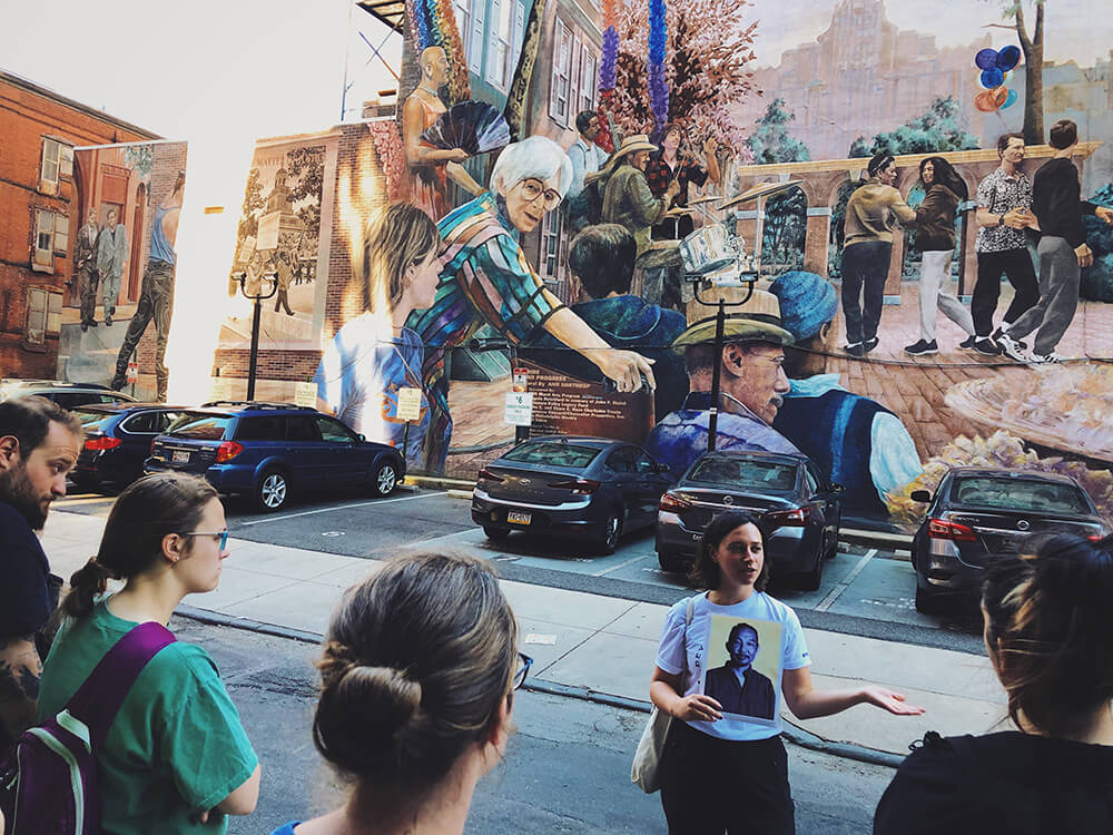 On the side of a building, a large colorful mural shows multiple women. In front of the mural, there are several parked cars. A tour guide stands in front of the mural, holding a portrait in her one hand as she addresses a group in front of her.