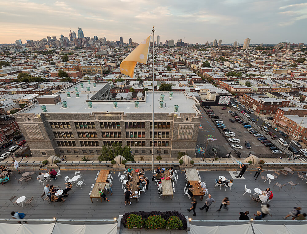 A rooftop bar with many people enjoying drinks at tables. A view of the city skyline is in the background.