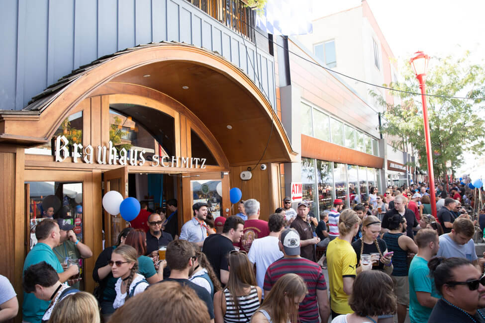 There is a large crowd of people standing outside on the sidewalk in front of a building with a sign that reads Brauhaus Schmitz. There are blue and white balloons tied to either side of the entrance. The doors are wide open.