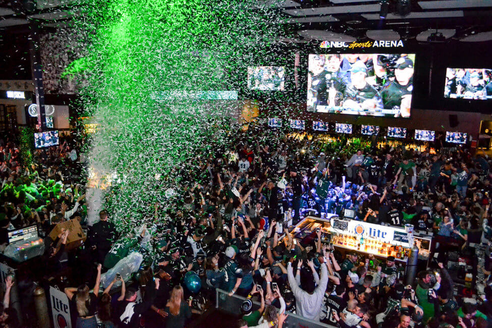 Cannons shoot green and white confetti into the air. There is a crowd of people celebrating, Many are wearing green jerseys and shirts. A large screen in the center of the indoor space shows a celebration a a coach is interviewed on camera. Fans throughout the space are seen cheering.