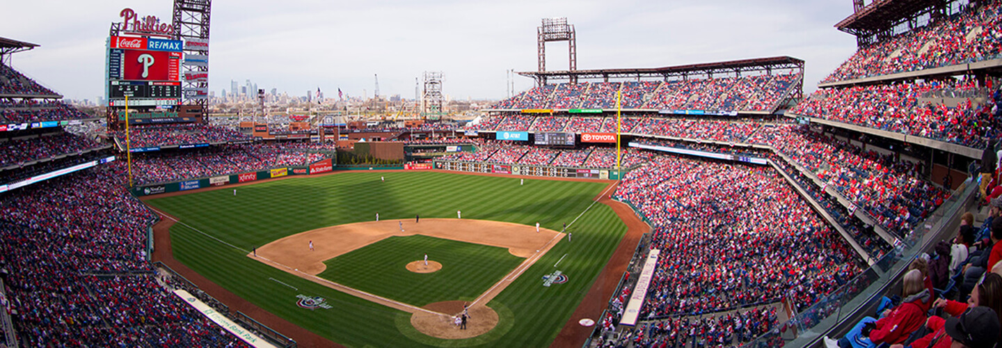 A baseball diamond is shown in the center. The stands surrounding the field are packed with fans wearing red, white, and blue, cheering on the Philadelphia Phillies. The sky is a light gray overhead.