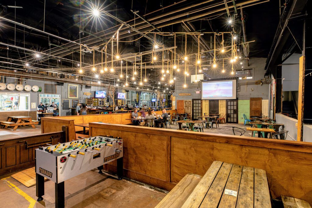 A large indoor space is shown. There are tables and chairs spread throughout, as well as picnic tables with benches attached. There are bright lights hanging from the ceiling. Along the wall to the left, there are multiple TV screens hanging above tables. There are people shown sitting around the tables.