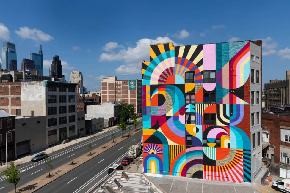 A colorful mural is shown on the side of the building. The sky above is blue with white clouds throughout. The street to the left of the mural is relatively clear, besides one car off to the far left.