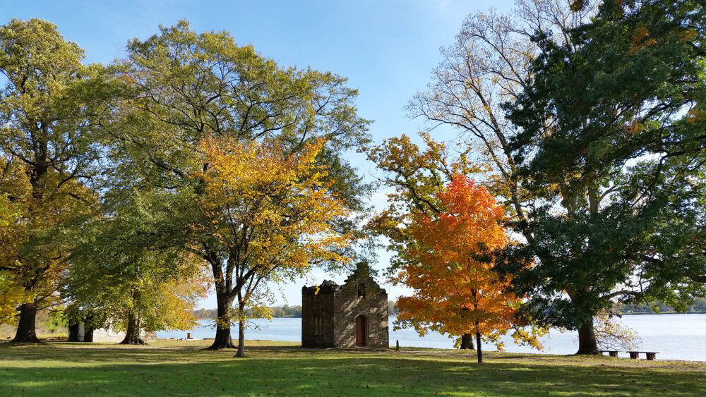 The view from a riverfront estate is shown. There is a structure in the middle. It is surrounded by colorful fall foliage on either side. The river is shown beyond it. The sky above is a bright blue. The grass is a bright green.