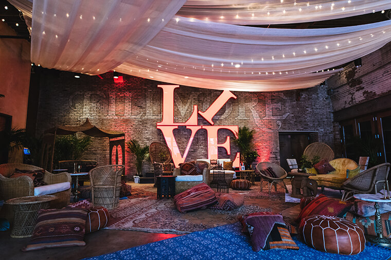 Seating is spread throughout the space. There are pieces of wicker furniture, bean bag chairs, and cushions. There is a large sign in the middle that reads LIVE in front of a brick wall that has FILLMORE written across it