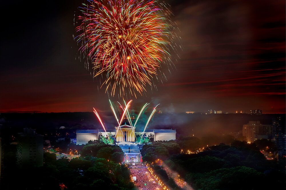 Fireworks shoot into the sky above the Philadelphia Museum of Art. The Benjamin Franklin Parkway is packed with people gazing up at the sky.