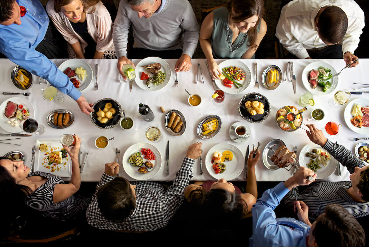 A bit of an aerial view of people sitting on both sides of a long table across the middle with plates of food as hands reach toward the items on the table