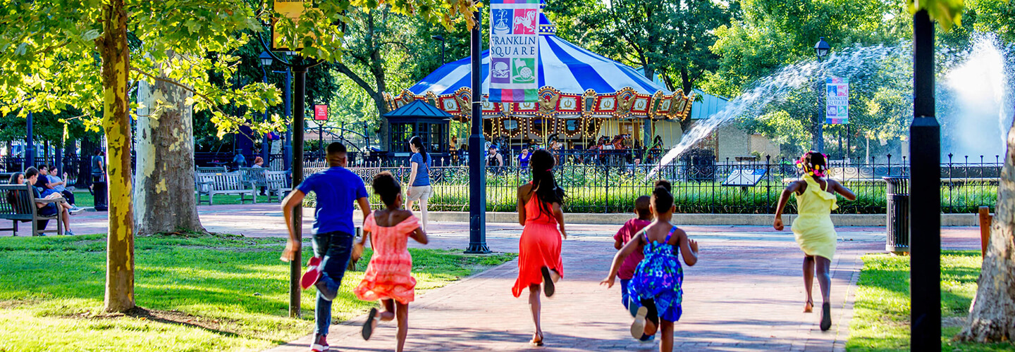 Five kids run through a park toward a colorful carousel