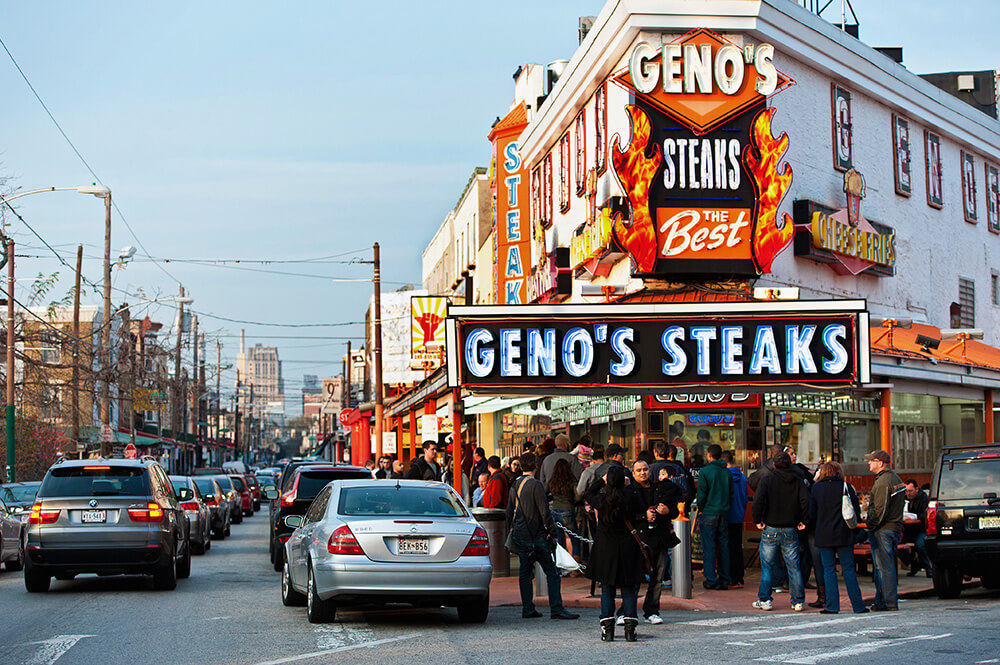 A steak shop on the corner of a city street is shown. There are people standing outside of the shop. There are bright orange and yellow neon signs and lights above them reading Geno's Steaks, The Best.