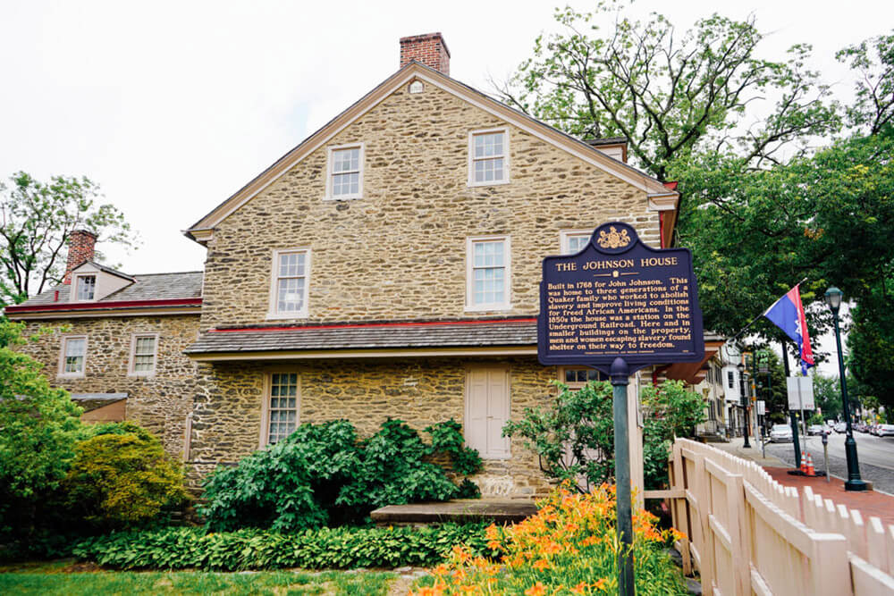 A blue historical marker sign reading The Johnson house is shown outside of the house. Green grass and bushes are in front of it, a tree is off to the right.