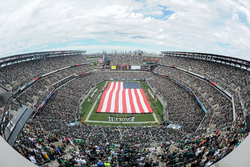 A large American flag is stretched out across the field at Lincoln Financial Field. The stands are packed with fans. The sky above the stadium is mainly white with clouds.