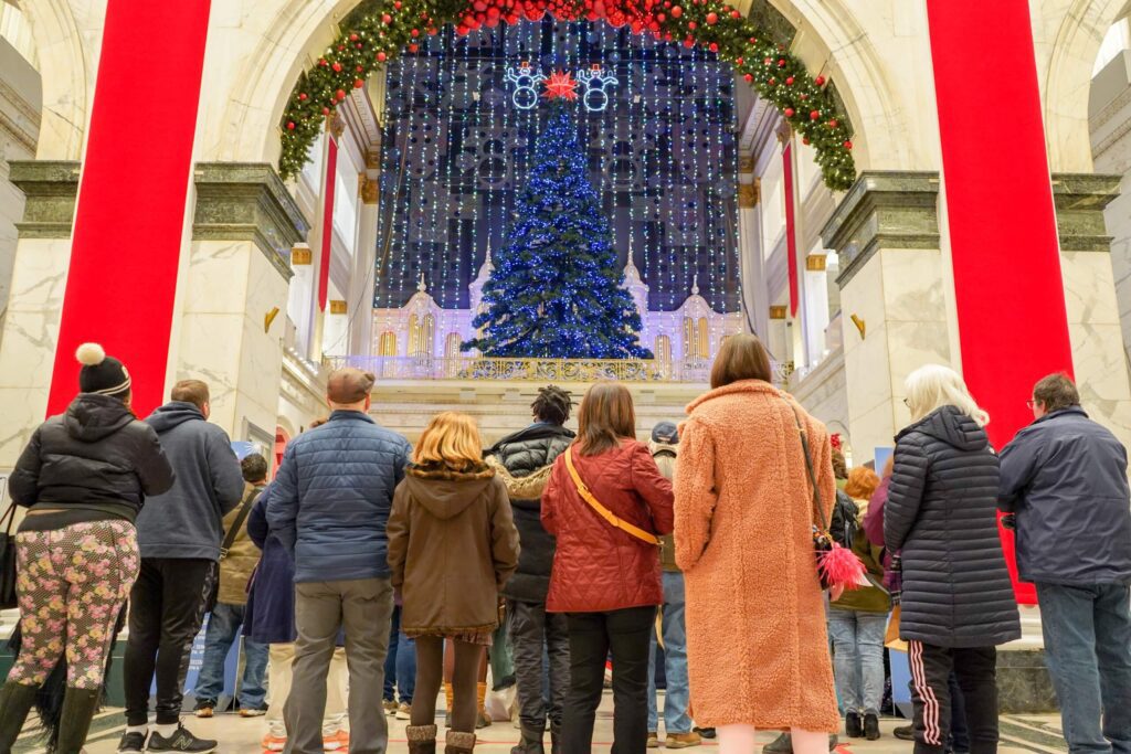 A group of people stand, looking up at a large Christmas tree surrounded by lights for the Macy's Christmas Light Show inside of a historic building. Two red decorative banners hang on either side