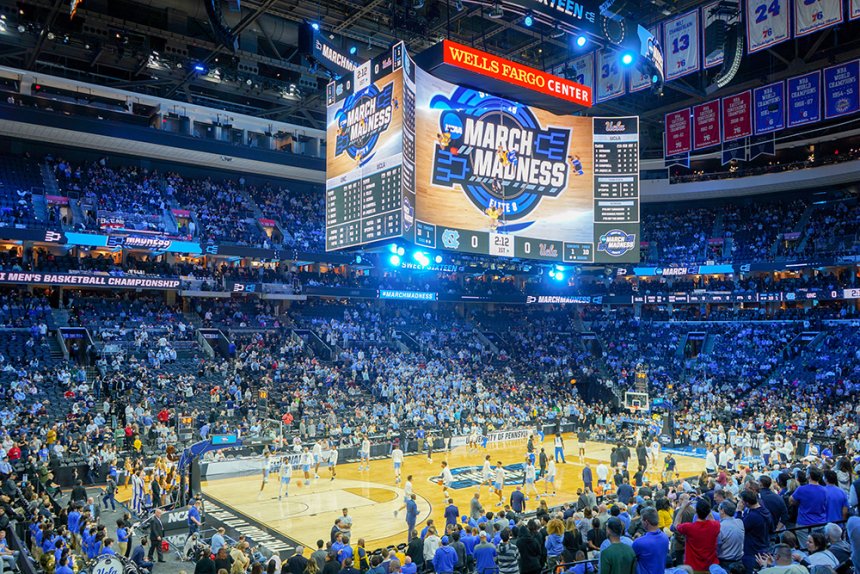 a basketball court is shown full of players. Coaches are lined up along the sides of the court. Fans are in the seats surrounding them. The jumbo tron hanging above the basketball court reads March Madness.