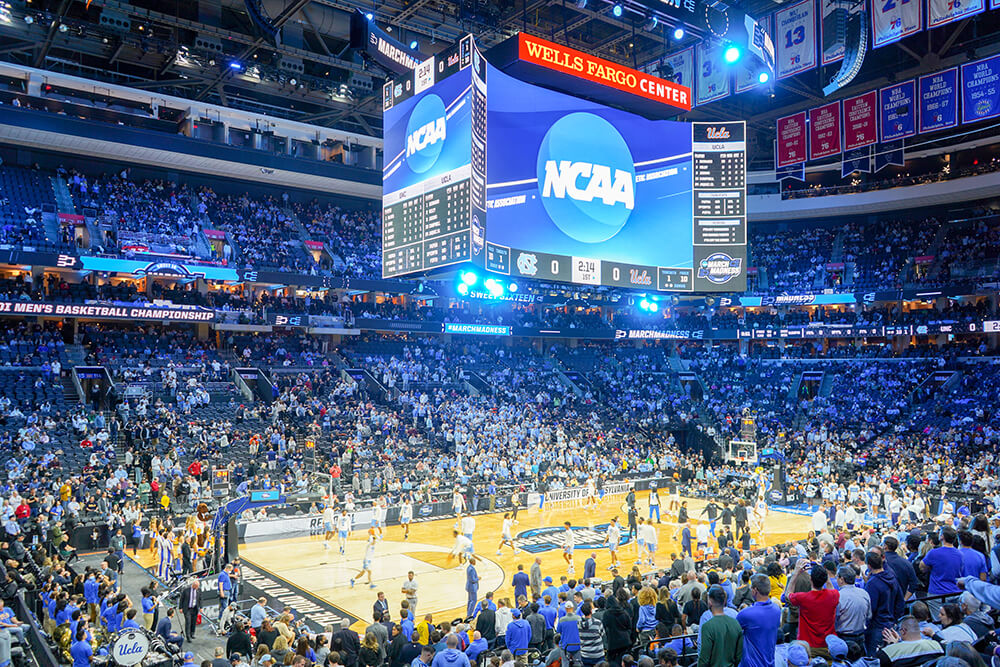 a basketball court is shown full of players. Coaches are lined up along the sides of the court. Fans are in the seats surrounding them. The jumbo tron hanging above the basketball court reads March Madness.