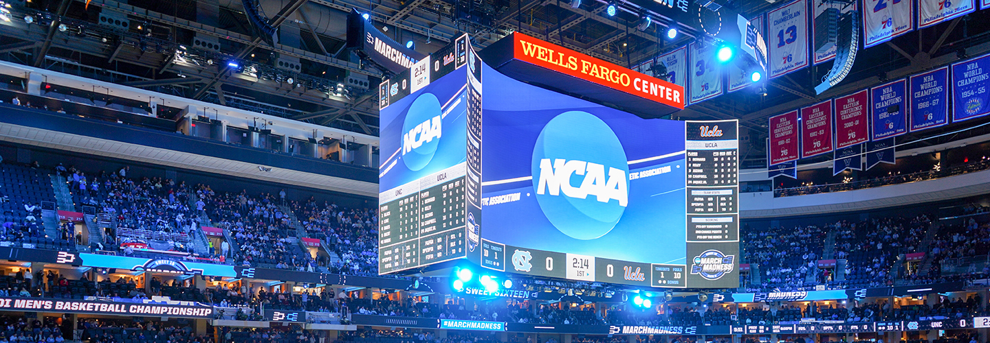 A jumbo tron in the center of an arena reads NCAA in white lettering against a blue background. Blue lights are casting a glow on the seats filled with fans.