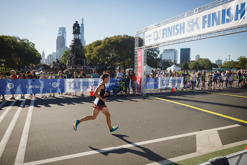 An athlete approaches the finish line after running the Philadelphia Distance Run. The sky is a light blue overhead. Spectators watch from behind the barriers set up along the course.