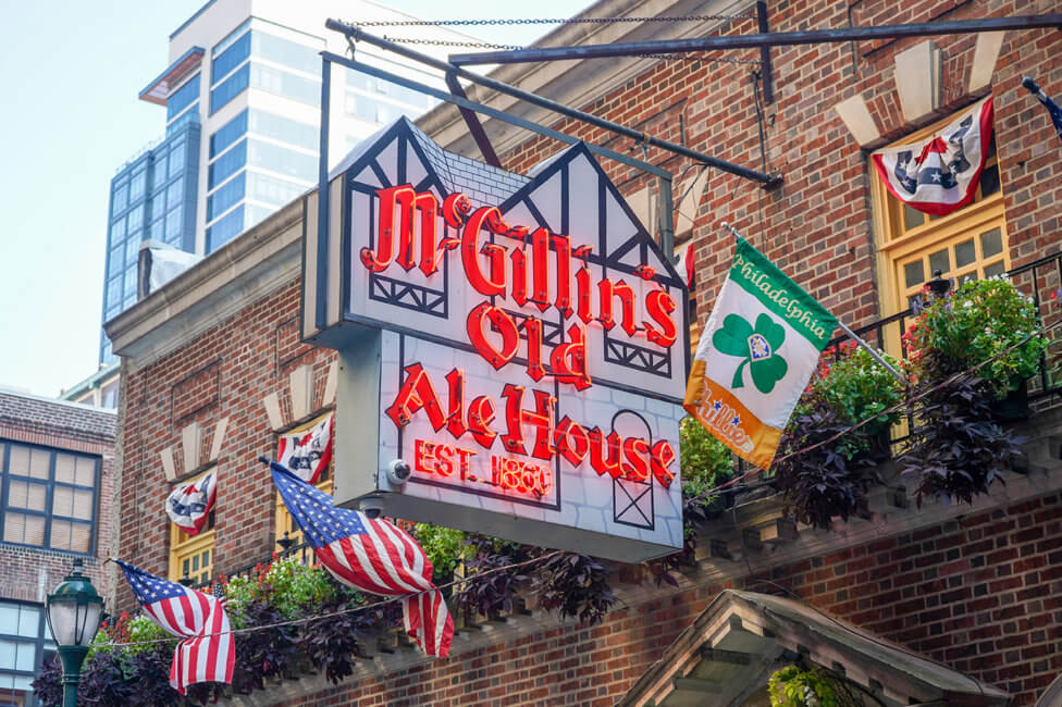A large sign hangs outside of a building. The sign reads McGillin's Olde Ale House Est. 1860 in bright reddish, orange letters. There are two American flags hanging off the building behind the sign. In front of the sign, there is a green, white, and orange flag that reads Philadelphia Phillies with a green shamrock in the center.