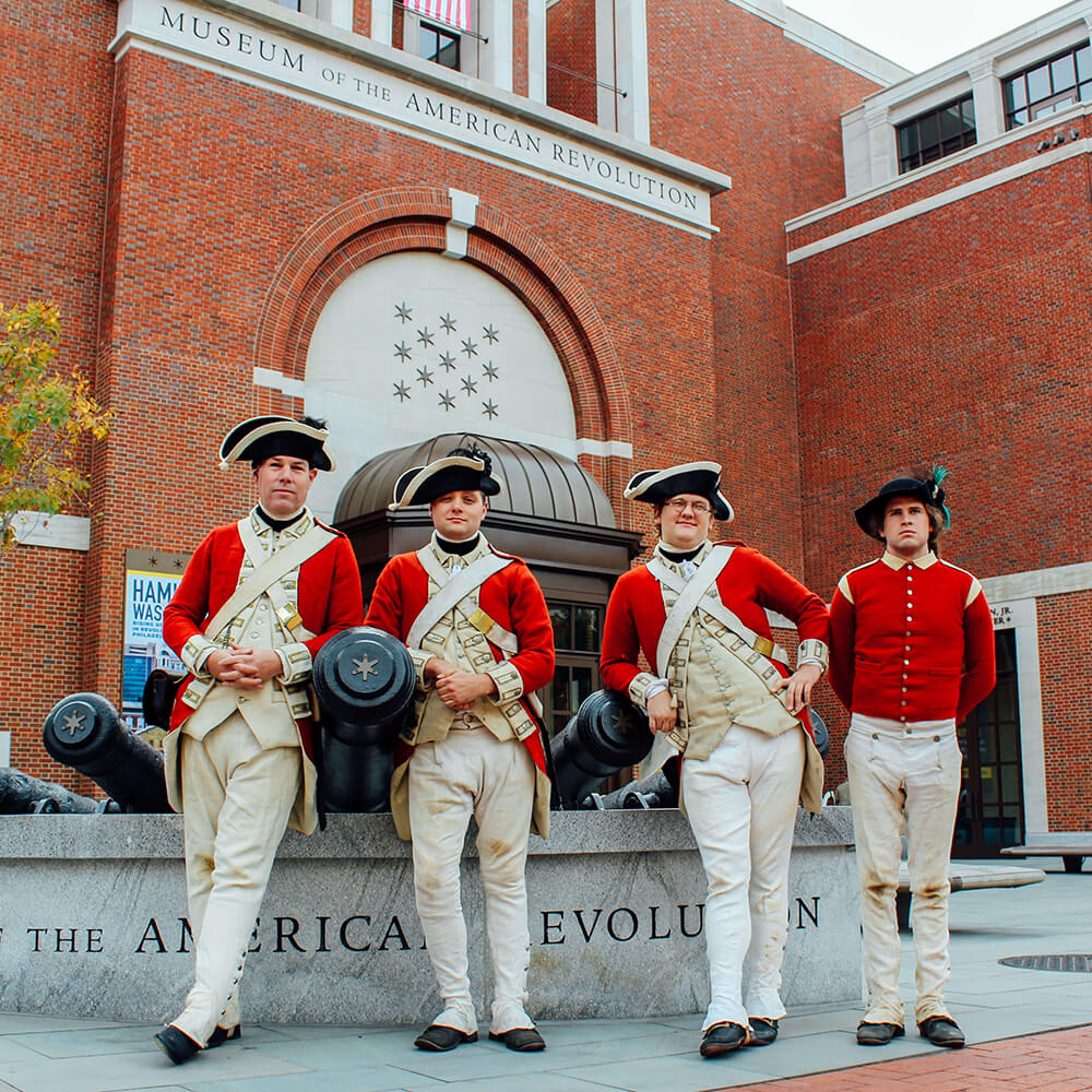 Four Revolutionary War reinactors stand in front of a cannon statue outside of a Museum
