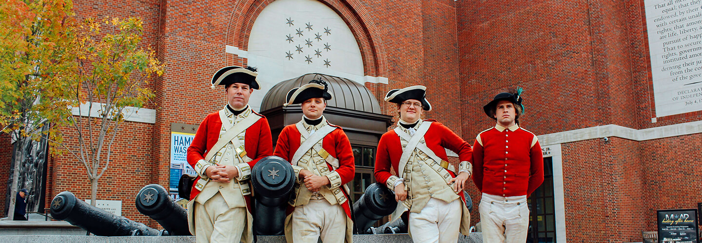 Four Revolutionary War reinactors stand in front of a cannon statue outside of a Museum
