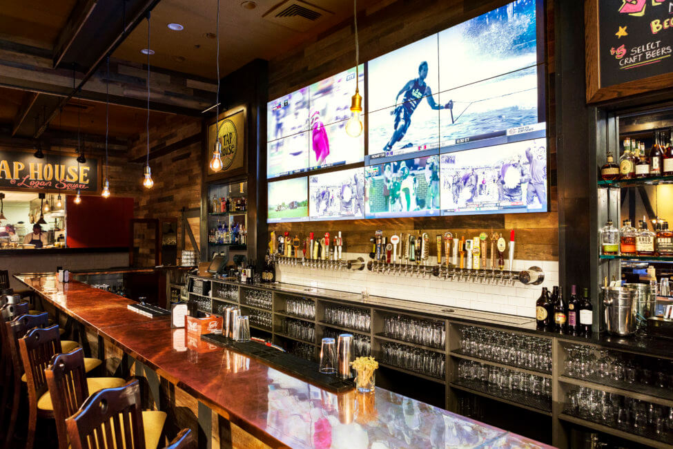 A large TV hangs above a bar that is fully stocked with glasses and bottles of alcohol. There is a large counter shown stretching across the front.