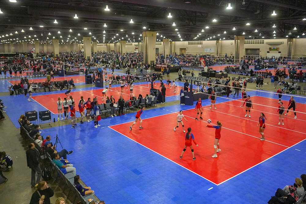 Inside of the Pennsylvania Convention Center, red and blue volleyball courts are set up throughout the space. Athletes compete on the court as coaches and spectators watch from the sidelines. Bright lights shine down from overhead.