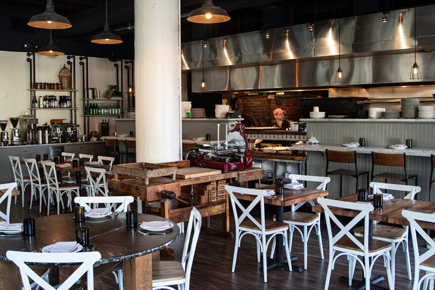 The interior of a restaurant is shown. There are white wooden chairs shown pushed up against tables. In the back, there is a cook in an open kitchen.