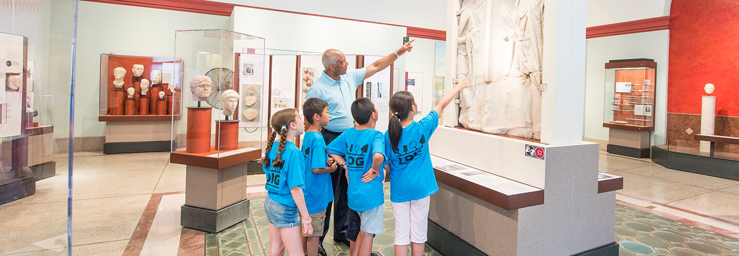 A group of students wearing blue t-shirts is shown with a chaperone. They stand looking at a display. A few of them are pointing.