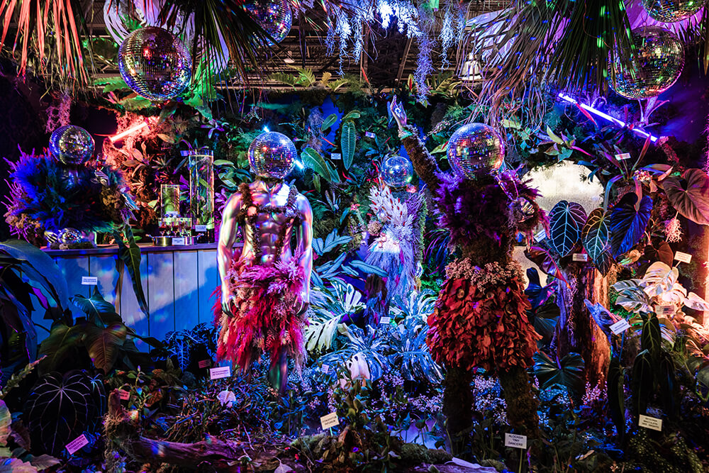 An elaborate and bright floral display at the Philadelphia Flower Show featuring mannequins wearing leis around their necks with discoballs as heads and floral skirts surrounded by exotic plants and flowers.