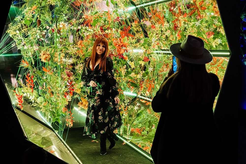 A woman with long red hair wearing a dark floral dress stands and poses for a picture in front of an elaborate floral display featuring green and orange plants. A silhouette of an individual wearing a hat and taking the woman's picture is shown off to the right.