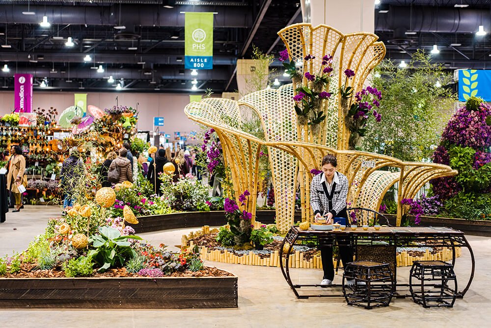 A woman is shown assembling items on a table in front of her off to the right in the frame. Behind her is a large, elaborate yellow structure, covered in purple and green flowers. To the left, there are raised garden beds holding beautiful floral arrangements. Off in the distance, attendees can be seen shopping at vendors set up throughout the space.